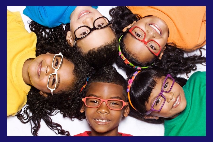 A group of five children with different skin tones lying on the ground in a star formation, smiling while wearing colorful shirts and glasses.