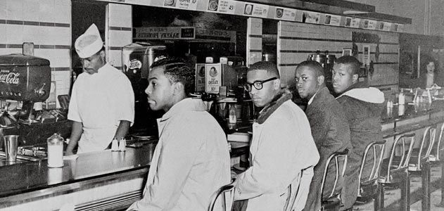 Black and white photo of four young men seated at a diner counter, looking forward. A man in a white chef's hat and apron is behind the counter preparing food.