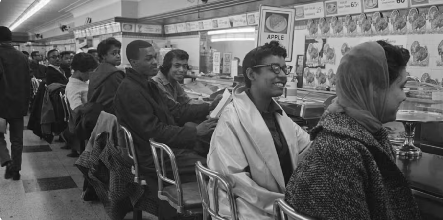 A group of people sitting at a lunch counter in a diner, engaging in conversation and smiling. The scene features individuals of diverse backgrounds, with a focus on a woman wearing glasses and a scarf. The decor includes menu signs on the wall.