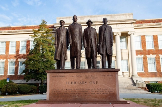 A sculpture of four men in formal attire standing together, commemorating the Greensboro Four civil rights activists, with an inscription reading 'FEBRUARY ONE' in front of a historic building.